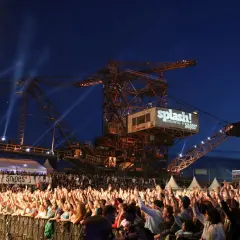 Große Menschenmenge jubelt bei einem Konzert vor einer riesigen Baggerschaufel mit dem Splash! Festival Line Up.