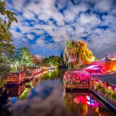 Bunte Lichter spiegeln sich im Wasser, Boote liegen am Ufer, ein belebtes Restaurant mit Menschen und eine Weide, die an die Reeperbahn in Berlin erinnert.