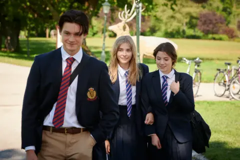 Drei Schüler in Schuluniformen gehen lächelnd durch einen Park. Ein Hirsch-Dekor im Hintergrund erinnert an die **serie gestern waren wir noch kinder**.