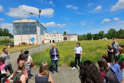 Gruppe bei einem **Festival im Osten** vor Industriegebäuden. Ein Mann erklärt etwas, während die Besucher zuhören.
