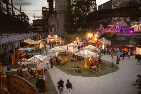 Besucher genießen das Ostrava Festival in einer ehemaligen Industrieanlage. Stände, Sitzbereiche und Lichter schaffen eine festliche Atmosphäre.