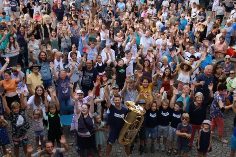 Große Menschenmenge winkt fröhlich bei Brackenheim Veranstaltungen. Ein Mann hält eine Tuba, Kinder und Erwachsene sind begeistert.