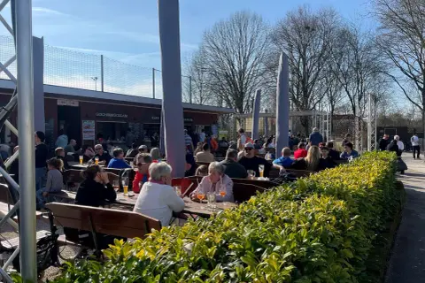Menschen genießen das sonnige Wetter im Carles Biergarten. Gäste sitzen an Holztischen, trinken und unterhalten sich.