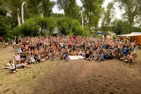 Große Gruppe feiert beim auerworld festival. Viele Menschen mit erhobenen Armen im Freien, umgeben von Bäumen und bunten Bannern.