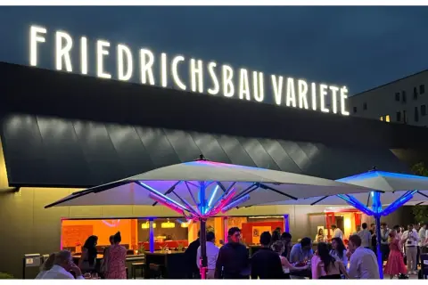 Leuchtende Buchstaben über dem Eingang des Varieté Friedrichsbau. Menschen versammeln sich unter beleuchteten Sonnenschirmen.