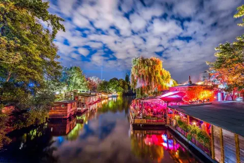 Bunte Lichter spiegeln sich im Wasser, Boote liegen am Ufer, ein belebtes Restaurant mit Menschen und eine Weide, die an die Reeperbahn in Berlin erinnert.