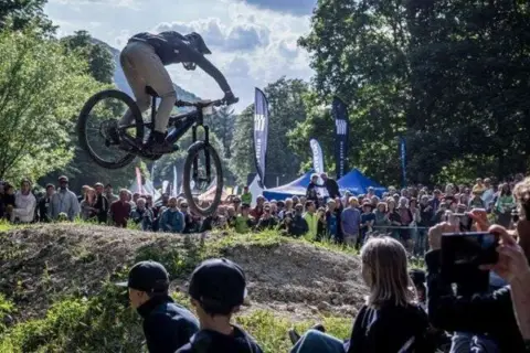 Mountainbiker springt bei einer Veranstaltung in Heubach heute über eine Schanze, während eine Menge zuschaut.