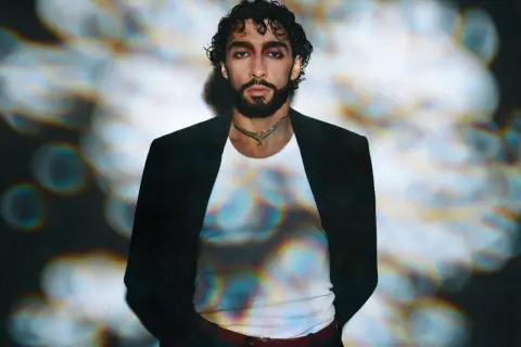 Sofiane Pamart in Berlin, mit dunklem Haar und Bart, trägt einen schwarzen Blazer über einem weißen Shirt. Bunte Lichtreflexe auf seinem Shirt.