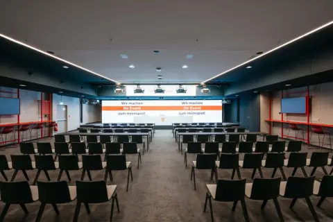 Moderne Konferenzräume im Kulturhaus Arena, bereit für Ihr Event. Reihenweise Stühle und eine große Leinwand mit der Botschaft "Wir machen Ihr Event zum Heimspiel!".