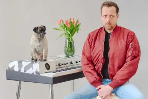 Ein Mops sitzt auf einem Keyboard, daneben ein Mann in roter Jacke. Eine Vase mit Tulpen steht zwischen ihnen. Eine gute Nachricht: Keine Gefahr!