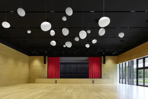 Moderne Aula mit Holzwänden, Bühne mit roten Vorhängen und vielen weißen Hängelampen. Die nobelgusch pfedelbach ist bereit für Veranstaltungen.
