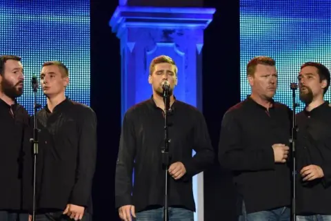 Fünf Männer in schwarzen Hemden singen auf einer Bühne. Sie präsentieren traditionelle kroatische Musik vor einem blauen, pixeligen Hintergrund.
