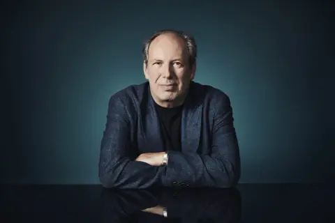 Hans Zimmer, der Meister der Filmmusik, sitzt mit verschränkten Armen vor einem dunklen Hintergrund. Sein Blick ist direkt auf die Kamera gerichtet.