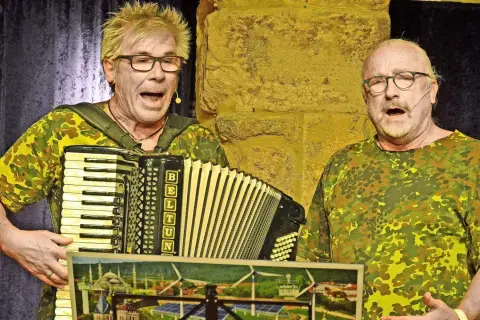 Zwei Männer im Tarnanzug singen und spielen Akkordeon beim **Kabarett der Galgenstricke**. Ein Bild mit Stadtansicht und Windrädern liegt vorn.