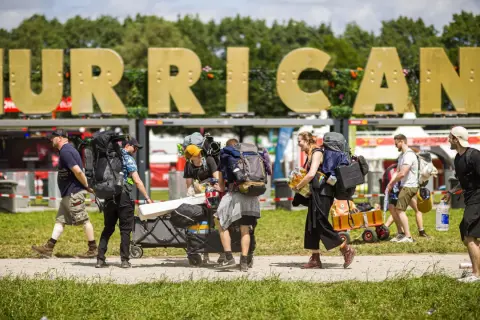 Besucher mit Rucksäcken und Gepäckwagen auf dem Hurricane Zeltplatz. Große goldene Buchstaben "HURRICANE" im Hintergrund.