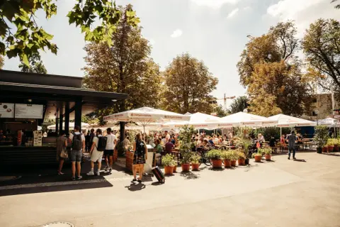 Sonniger Biergarten im Schlossgarten mit vielen Gästen unter schattenspendenden Schirmen.