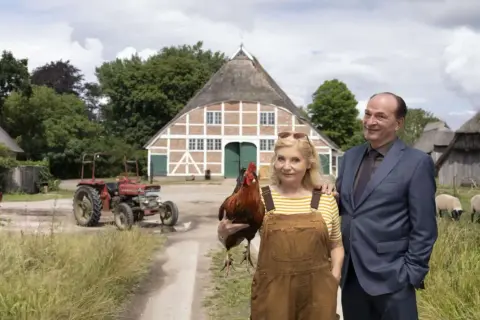 Ein Mann im Anzug und eine Frau mit Latzhose und Huhn stehen vor einem Bauernhaus. Im **weiten Land** grasen Schafe, ein Traktor steht bereit.