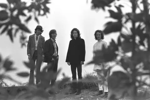 Die Mitglieder von The Doors stehen in einer Reihe, umgeben von Ästen und Blättern.