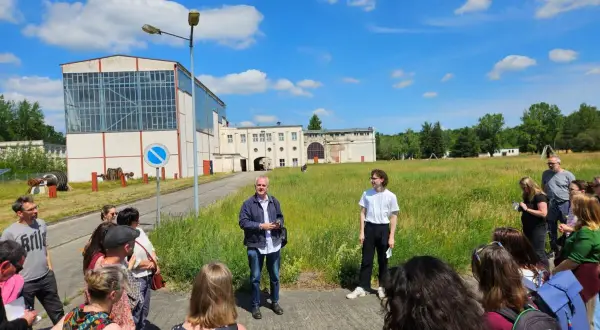 Gruppe bei einem **Festival im Osten** vor Industriegebäuden. Ein Mann erklärt etwas, während die Besucher zuhören.