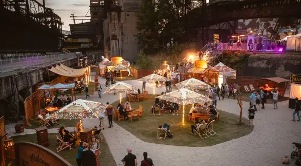 Besucher genießen das Ostrava Festival in einer ehemaligen Industrieanlage. Stände, Sitzbereiche und Lichter schaffen eine festliche Atmosphäre.