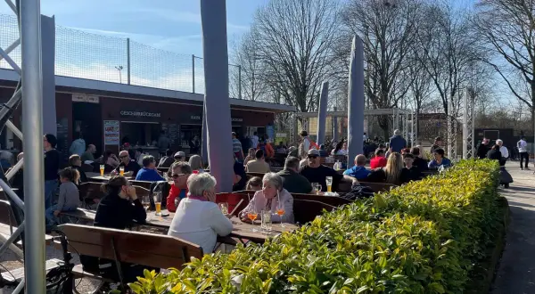 Menschen genießen das sonnige Wetter im Carles Biergarten. Gäste sitzen an Holztischen, trinken und unterhalten sich.