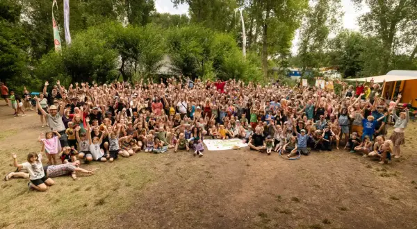 Große Gruppe feiert beim auerworld festival. Viele Menschen mit erhobenen Armen im Freien, umgeben von Bäumen und bunten Bannern.
