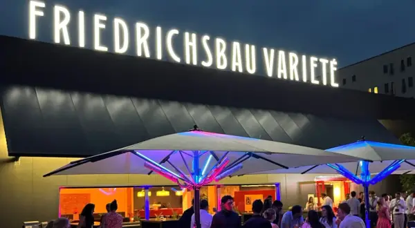 Leuchtende Buchstaben über dem Eingang des Varieté Friedrichsbau. Menschen versammeln sich unter beleuchteten Sonnenschirmen.