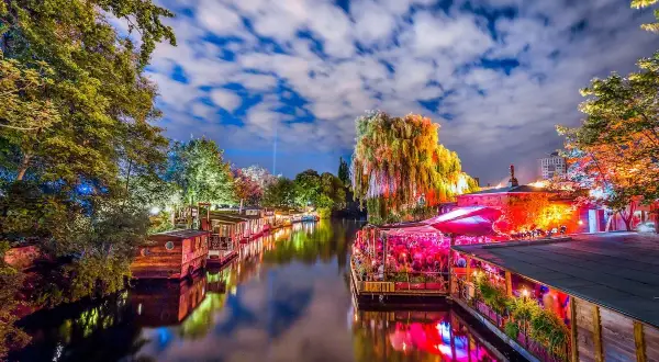 Bunte Lichter spiegeln sich im Wasser, Boote liegen am Ufer, ein belebtes Restaurant mit Menschen und eine Weide, die an die Reeperbahn in Berlin erinnert.