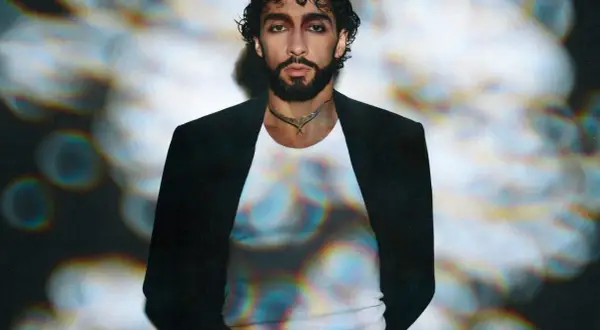 Sofiane Pamart in Berlin, mit dunklem Haar und Bart, trägt einen schwarzen Blazer über einem weißen Shirt. Bunte Lichtreflexe auf seinem Shirt.