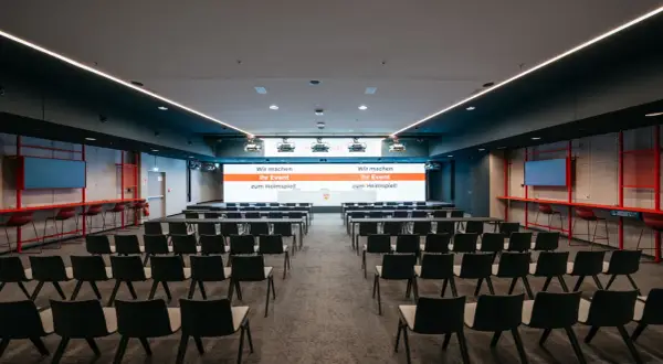 Moderne Konferenzräume im Kulturhaus Arena, bereit für Ihr Event. Reihenweise Stühle und eine große Leinwand mit der Botschaft "Wir machen Ihr Event zum Heimspiel!".