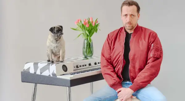 Ein Mops sitzt auf einem Keyboard, daneben ein Mann in roter Jacke. Eine Vase mit Tulpen steht zwischen ihnen. Eine gute Nachricht: Keine Gefahr!
