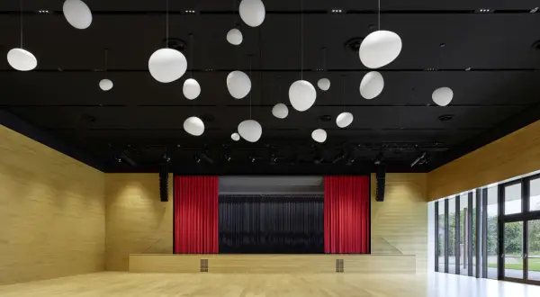 Moderne Aula mit Holzwänden, Bühne mit roten Vorhängen und vielen weißen Hängelampen. Die nobelgusch pfedelbach ist bereit für Veranstaltungen.