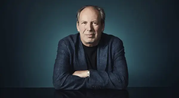 Hans Zimmer, der Meister der Filmmusik, sitzt mit verschränkten Armen vor einem dunklen Hintergrund. Sein Blick ist direkt auf die Kamera gerichtet.