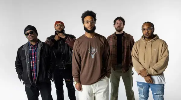 Fünf Männer stehen vor weißem Hintergrund. Einer trägt ein **butcher brown** Langarmshirt mit einem Hirschgeweih-Symbol.