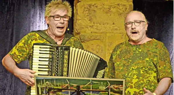 Zwei Männer im Tarnanzug singen und spielen Akkordeon beim **Kabarett der Galgenstricke**. Ein Bild mit Stadtansicht und Windrädern liegt vorn.