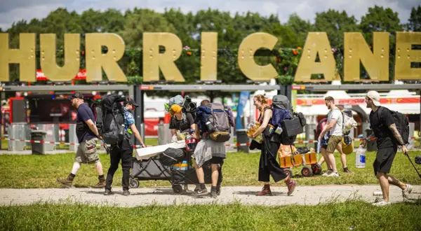 Besucher mit Rucksäcken und Gepäckwagen auf dem Hurricane Zeltplatz. Große goldene Buchstaben "HURRICANE" im Hintergrund.