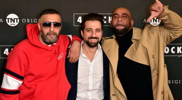 Drei Männer posieren vor einem Banner mit "4 Blocks". Einer trägt eine rote Kapuzenjacke, der andere einen beigen Trenchcoat.