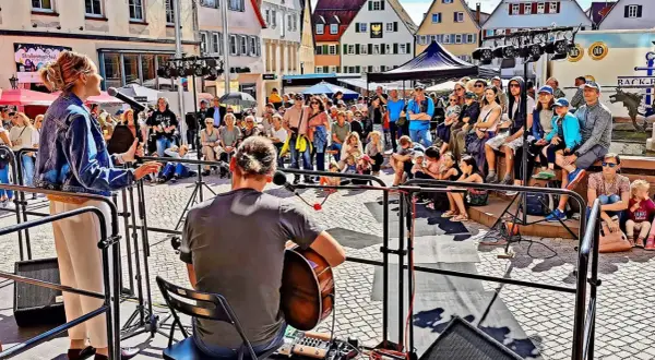Ein Musiker spielt Gitarre, während eine Sängerin auf einer Bühne steht. Viele Menschen sitzen und stehen davor, um die **Veranstaltung weil der Stadt** zu genießen.