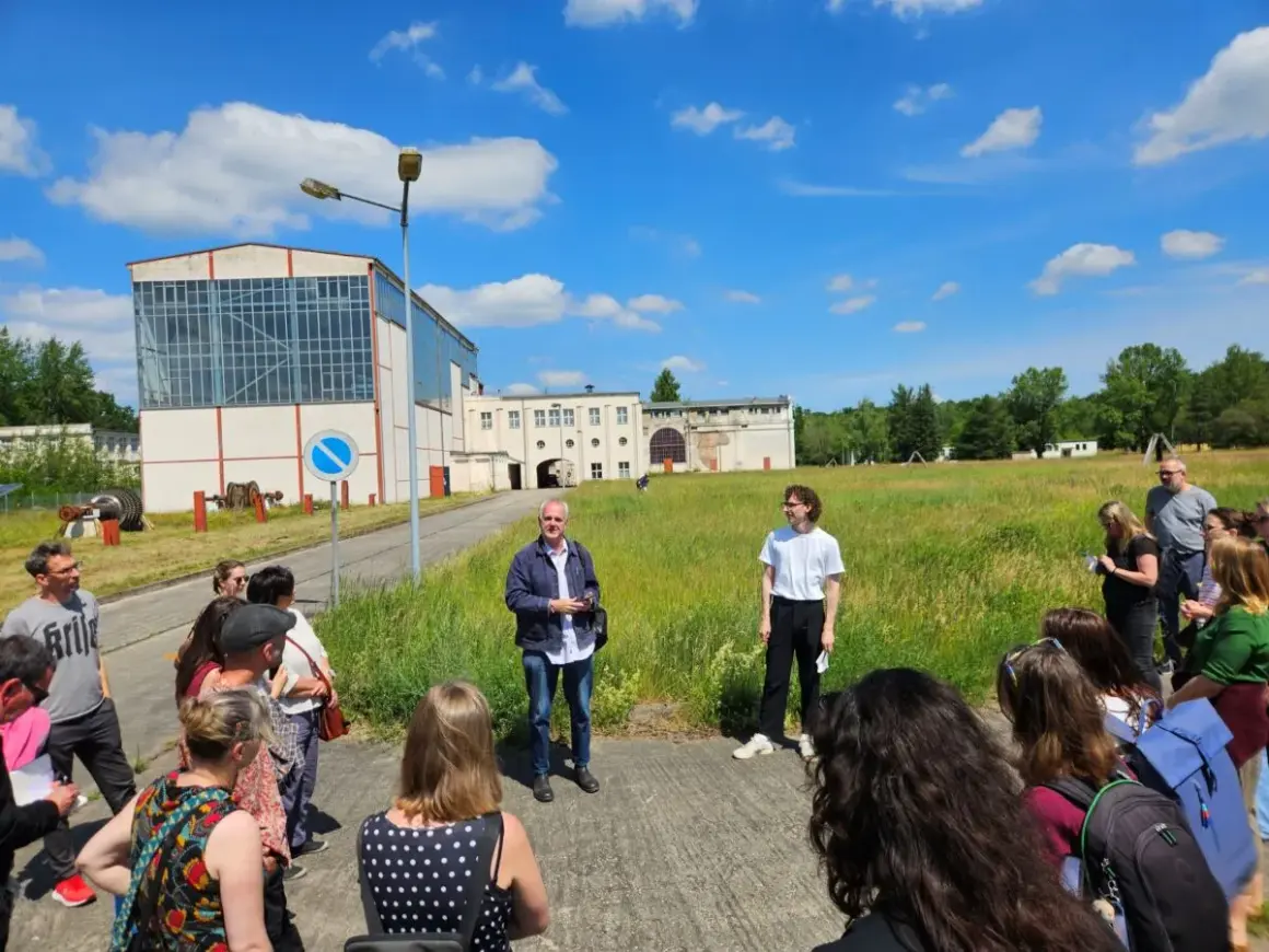 Gruppe bei einem **Festival im Osten** vor Industriegebäuden. Ein Mann erklärt etwas, während die Besucher zuhören.