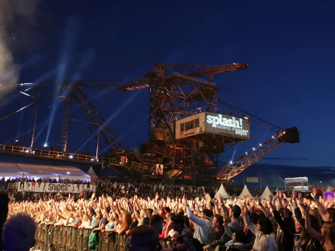 Große Menschenmenge jubelt bei einem Konzert vor einer riesigen Baggerschaufel mit dem Splash! Festival Line Up.