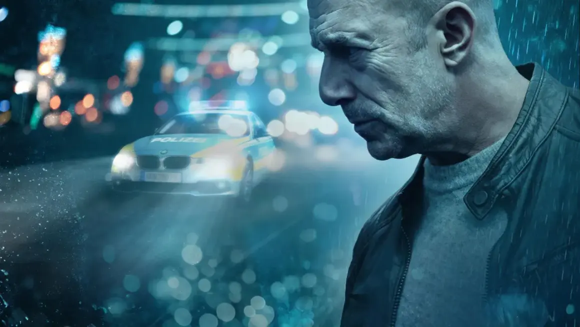 Heino Ferch im düsteren Krimi-Setting. Ein Polizeiauto mit Blaulicht im Regen, die Stimmung ist angespannt. Mehr im Krimi in der Mediathek.