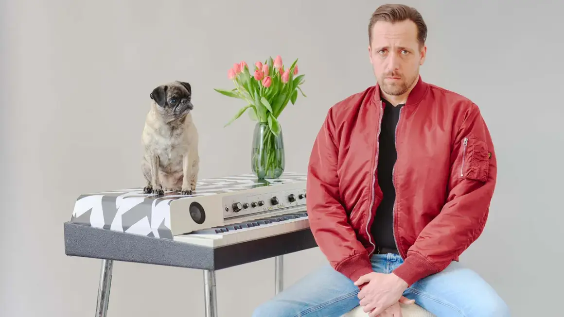 Ein Mops sitzt auf einem Keyboard, daneben ein Mann in roter Jacke. Eine Vase mit Tulpen steht zwischen ihnen. Eine gute Nachricht: Keine Gefahr!