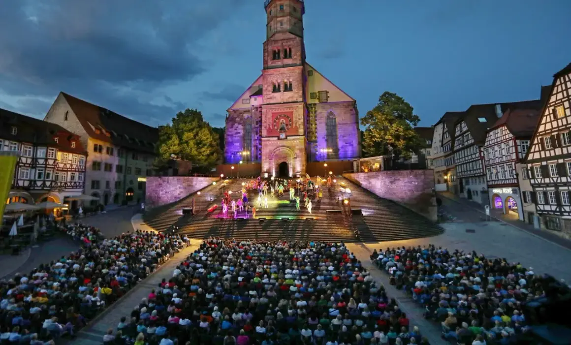 Bühnenaufführung vor historischer Kulisse in Hohenlohe. Ein großes Publikum verfolgt gespannt die Darbietung auf illuminierten Treppen vor einer Kirche.