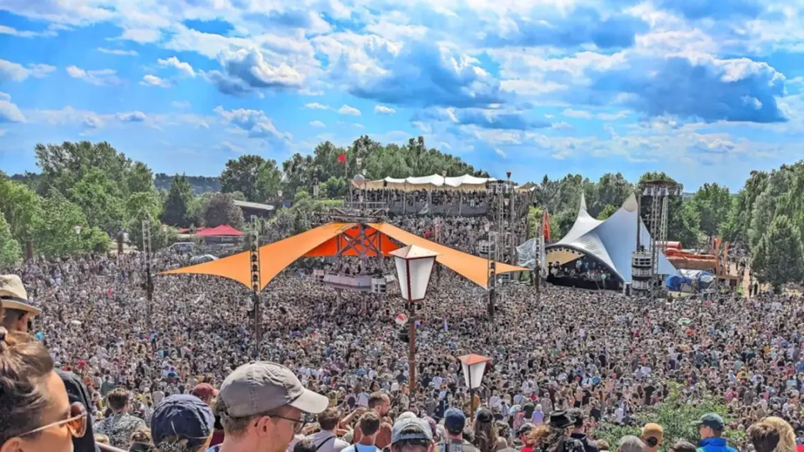 Tausende feiern auf dem Fusion Festival Ort unter blauem Himmel. Bunte Zelte und Bühnen prägen die Szenerie.