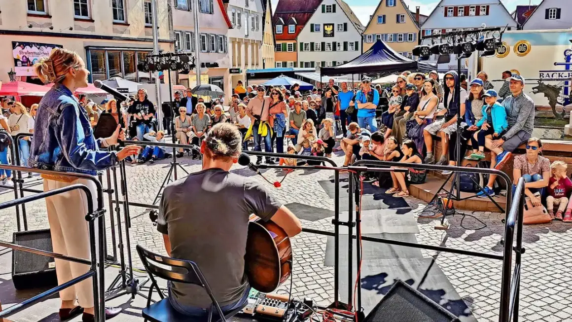 Ein Musiker spielt Gitarre, während eine Sängerin auf einer Bühne steht. Viele Menschen sitzen und stehen davor, um die **Veranstaltung weil der Stadt** zu genießen.