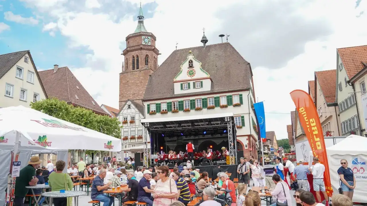 Festliche Atmosph&auml;re bei einem Veranstaltung weil der Stadt. Eine Blasmusik spielt auf einer B&uuml;hne, w&auml;hrend Menschen an Tischen sitzen und das bayerische Flair genie&szlig;en.