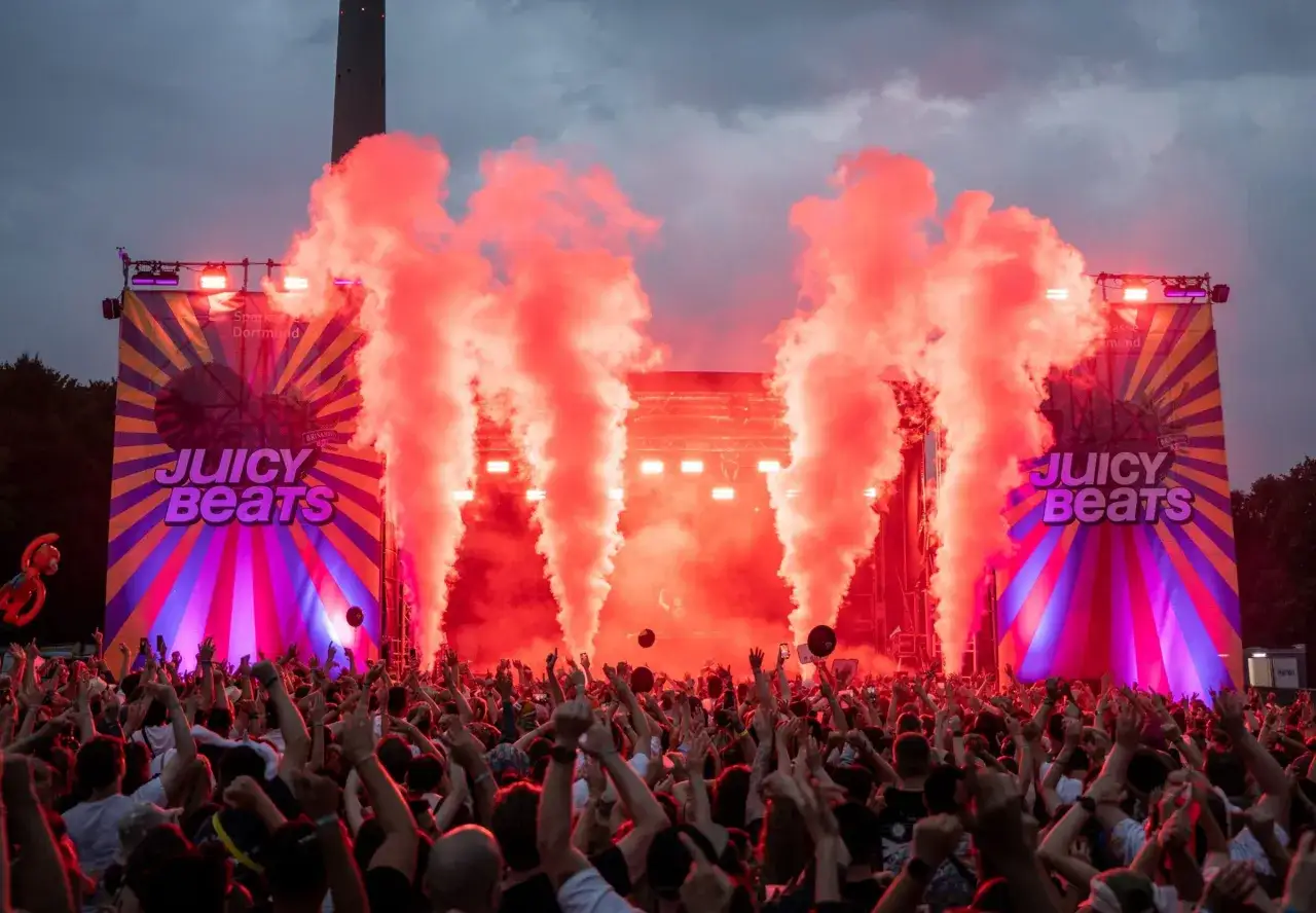 Rote Rauchschwaden steigen auf bei Juicy Beats 2025 in Dortmund. Eine jubelnde Menge feiert die Musik unter einem dramatischen Himmel.
