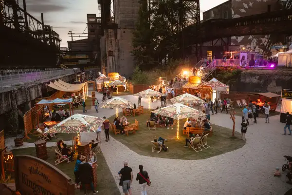 Besucher genießen das Ostrava Festival in einer ehemaligen Industrieanlage. Stände, Sitzbereiche und Lichter schaffen eine festliche Atmosphäre.