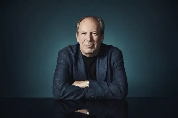 Hans Zimmer, der Meister der Filmmusik, sitzt mit verschränkten Armen vor einem dunklen Hintergrund. Sein Blick ist direkt auf die Kamera gerichtet.